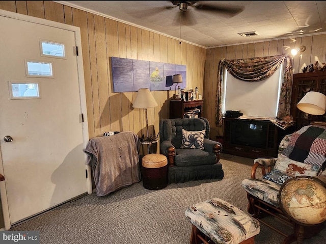 carpeted living area featuring wooden walls, visible vents, crown molding, and a ceiling fan