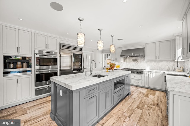 kitchen with a warming drawer, gray cabinets, a sink, wall chimney exhaust hood, and built in appliances