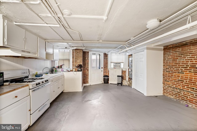 kitchen featuring under cabinet range hood, white gas range oven, white cabinetry, brick wall, and concrete flooring