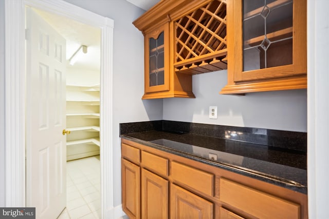 kitchen with light tile patterned flooring, glass insert cabinets, brown cabinetry, and dark stone countertops