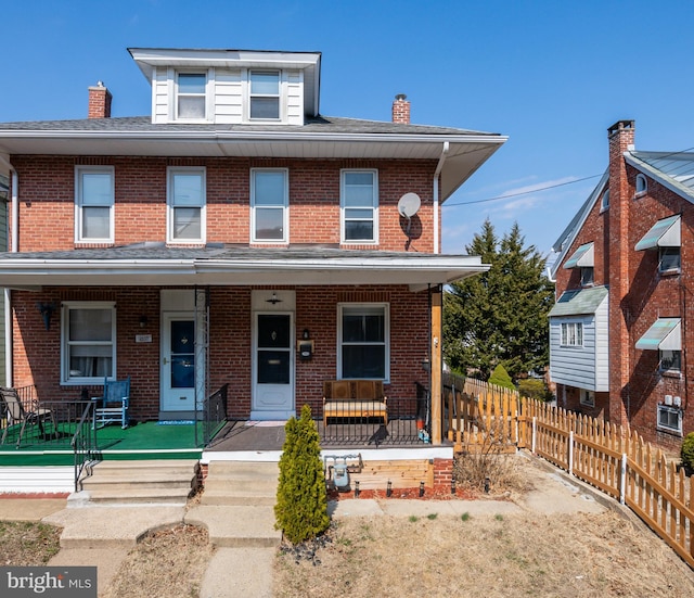 american foursquare style home featuring a porch, fence, roof with shingles, brick siding, and a chimney