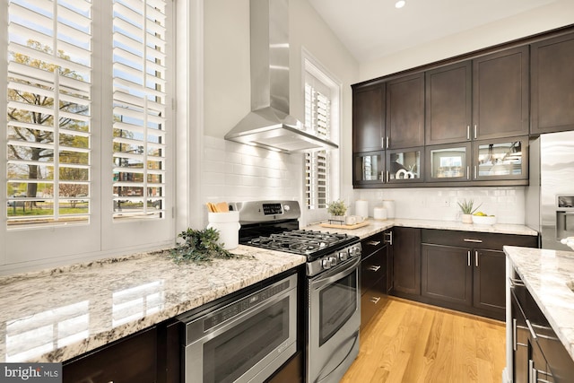 kitchen with wall chimney exhaust hood, dark brown cabinetry, a wealth of natural light, and appliances with stainless steel finishes