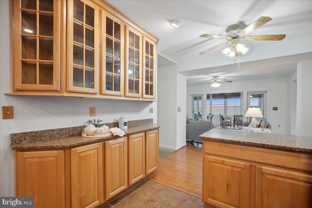 kitchen featuring stone countertops, open floor plan, glass insert cabinets, baseboards, and ceiling fan