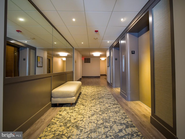 corridor featuring wood finished floors, baseboards, elevator, recessed lighting, and a paneled ceiling