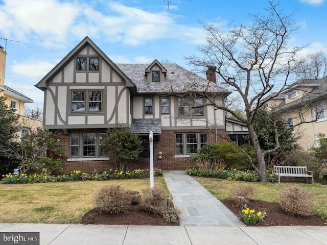 tudor home featuring stucco siding, brick siding, a chimney, and a front lawn