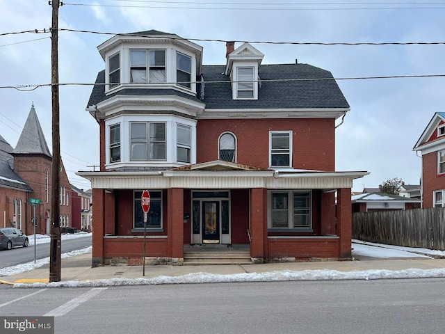 view of front facade featuring brick siding, covered porch, roof with shingles, and fence