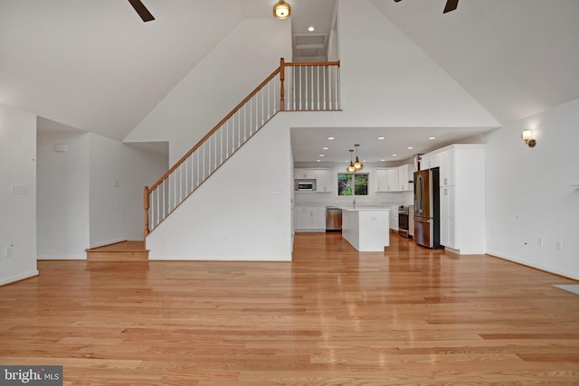 unfurnished living room with stairway, light wood-style flooring, high vaulted ceiling, and ceiling fan