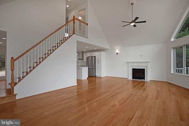 unfurnished living room with a ceiling fan, light wood finished floors, a fireplace with flush hearth, stairs, and a towering ceiling