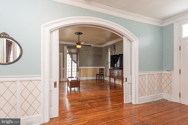 corridor with a healthy amount of sunlight, a wainscoted wall, ornamental molding, wood finished floors, and arched walkways