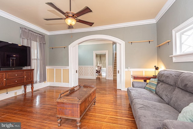 living room with a ceiling fan, wood finished floors, arched walkways, wainscoting, and crown molding