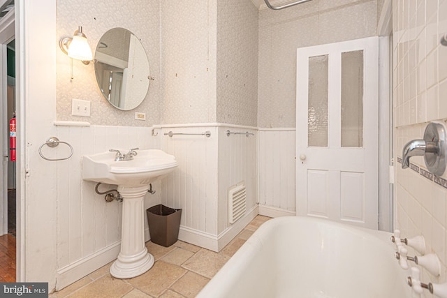 bathroom with stone tile floors, wainscoting, a washtub, and wallpapered walls