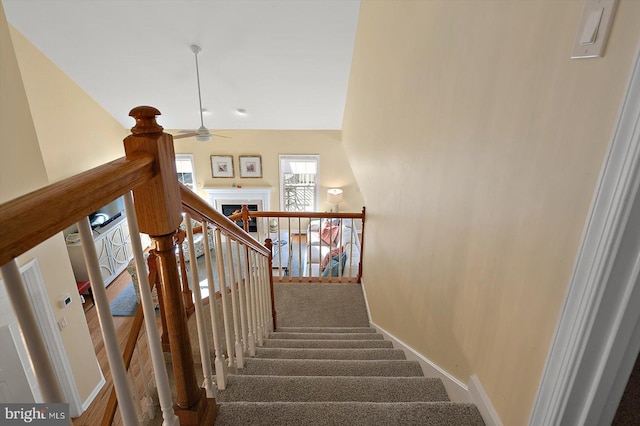 staircase with baseboards, a warm lit fireplace, and ceiling fan