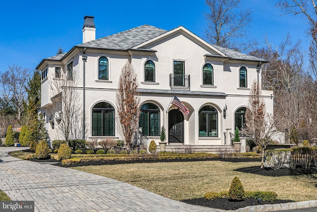 french country style house with stucco siding, a front yard, a balcony, and a chimney