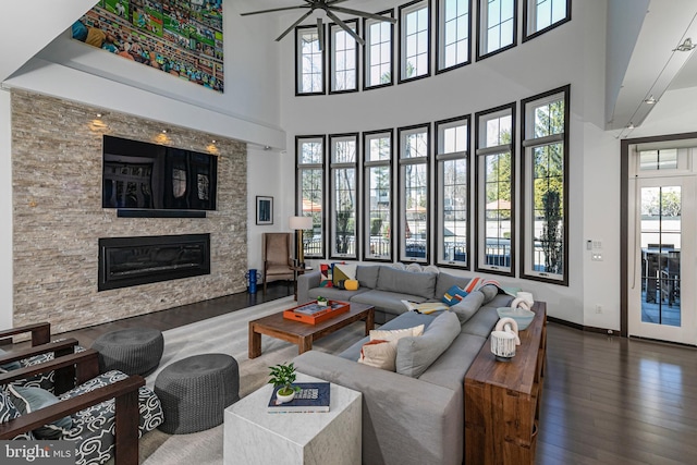 living area with baseboards, ceiling fan, a stone fireplace, a high ceiling, and wood finished floors