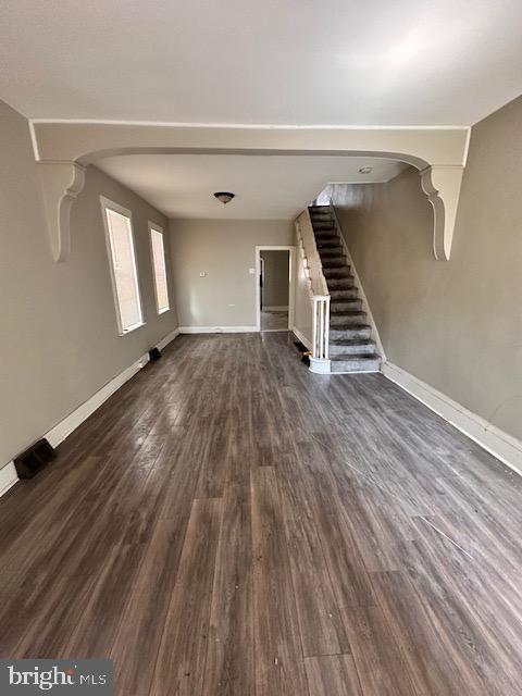 unfurnished living room with stairs, arched walkways, baseboards, and dark wood-style flooring