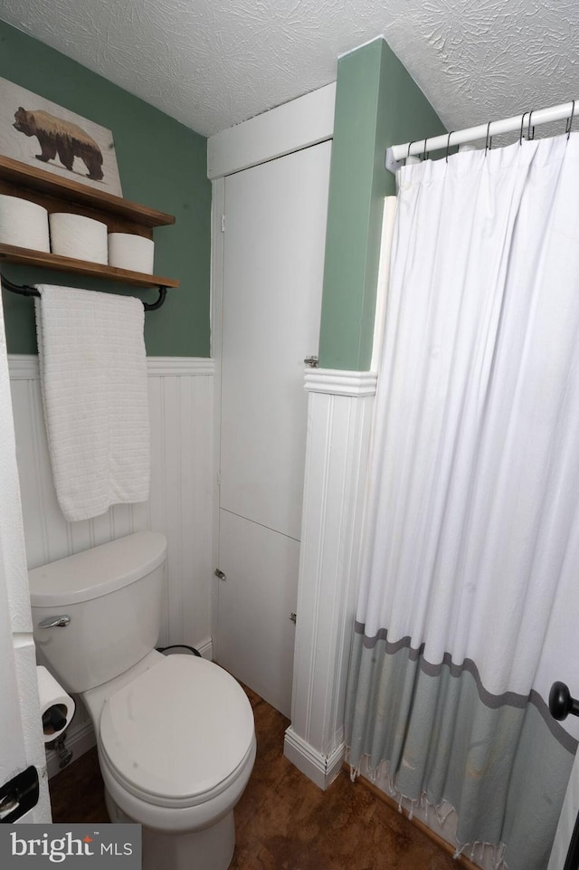 bathroom featuring a shower with shower curtain, toilet, a wainscoted wall, and a textured ceiling