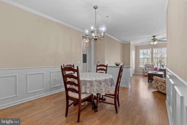 dining space with light wood finished floors, a wainscoted wall, ceiling fan with notable chandelier, and ornamental molding