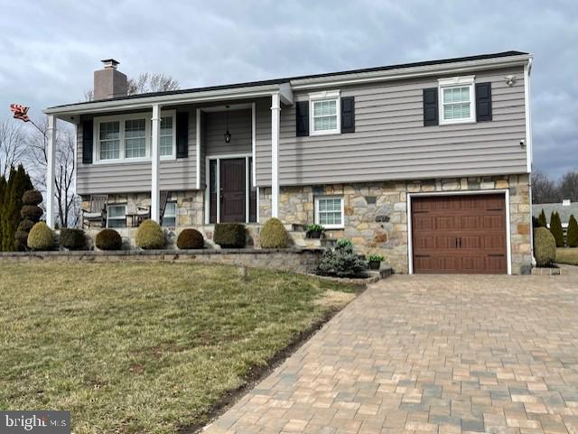 bi-level home with a front yard, decorative driveway, stone siding, and a chimney