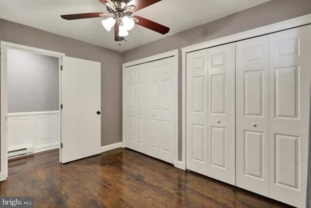 unfurnished bedroom featuring a ceiling fan, a wainscoted wall, a baseboard radiator, dark wood finished floors, and two closets