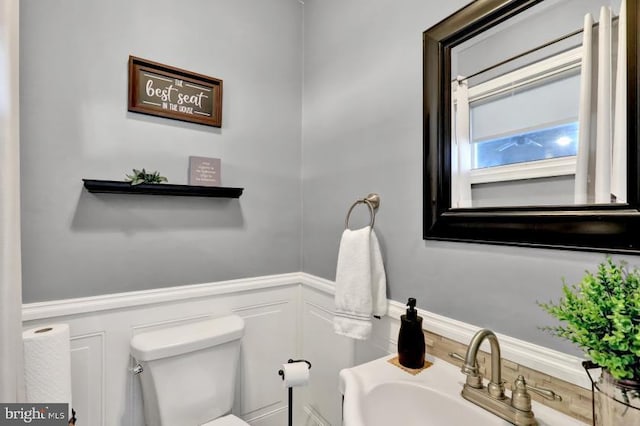 bathroom featuring a sink, a decorative wall, toilet, and wainscoting