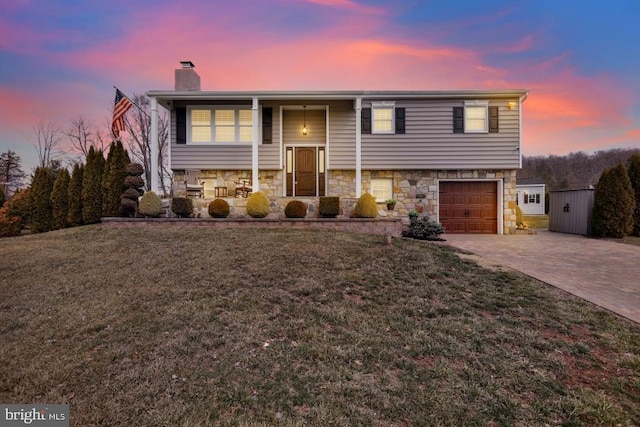 raised ranch with a yard, an attached garage, a chimney, stone siding, and decorative driveway