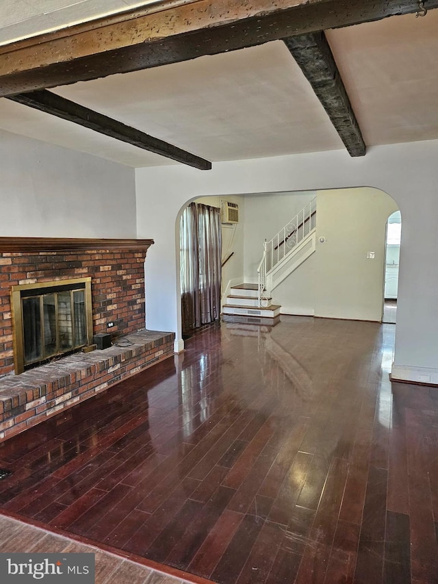 unfurnished living room featuring a brick fireplace, stairs, beam ceiling, wood finished floors, and arched walkways