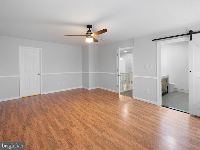 unfurnished bedroom with a ceiling fan, wood finished floors, baseboards, ensuite bathroom, and a barn door