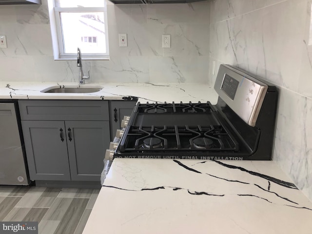 kitchen with light stone counters, gray cabinetry, a sink, appliances with stainless steel finishes, and backsplash