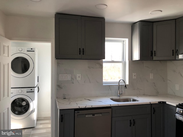 kitchen featuring gray cabinetry, a sink, light stone counters, stainless steel dishwasher, and stacked washer / dryer