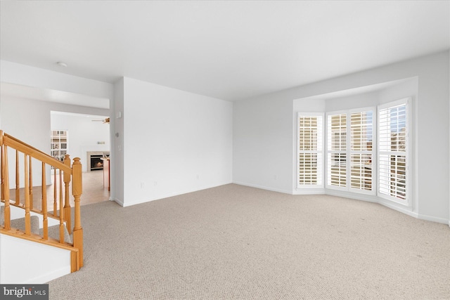 unfurnished room with stairway, baseboards, a lit fireplace, ceiling fan, and carpet flooring