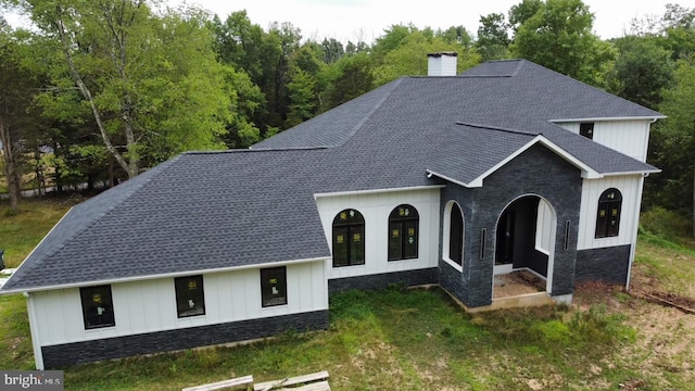 view of front facade with a front yard, roof with shingles, a chimney, stone siding, and board and batten siding