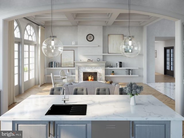 kitchen with light stone counters, a kitchen island with sink, coffered ceiling, and a lit fireplace