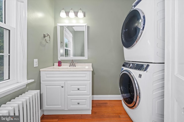 clothes washing area with wood finished floors, baseboards, radiator heating unit, a sink, and stacked washer and clothes dryer