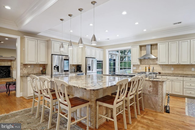 kitchen featuring light wood-type flooring, appliances with stainless steel finishes, wall chimney exhaust hood, and a large island with sink