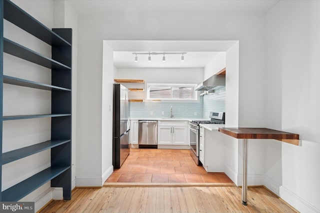 kitchen with open shelves, a sink, light wood-style floors, appliances with stainless steel finishes, and wall chimney range hood