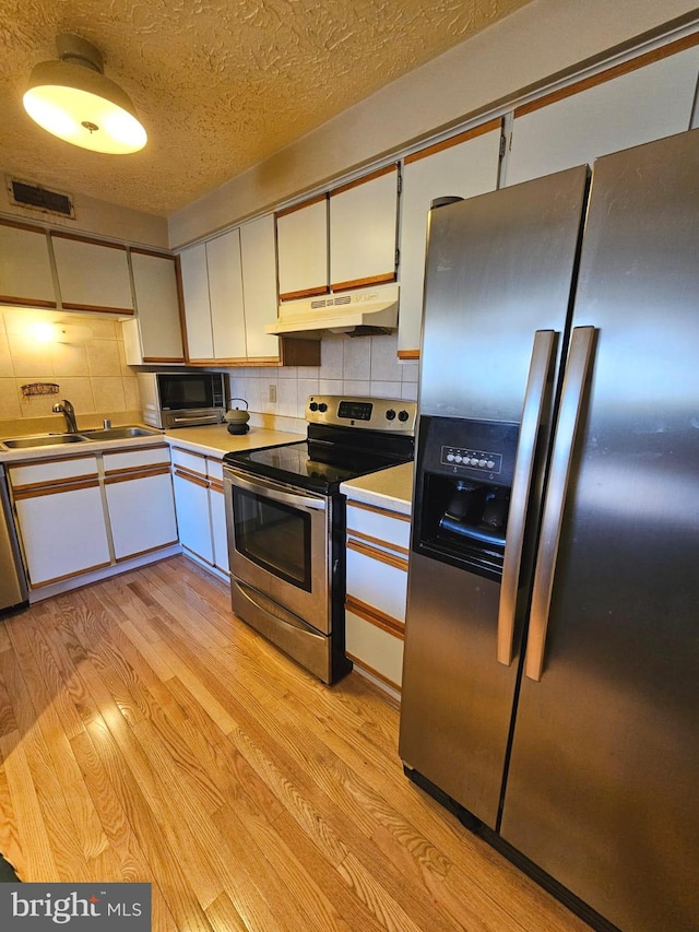 kitchen with a sink, stainless steel appliances, light wood-style floors, under cabinet range hood, and tasteful backsplash