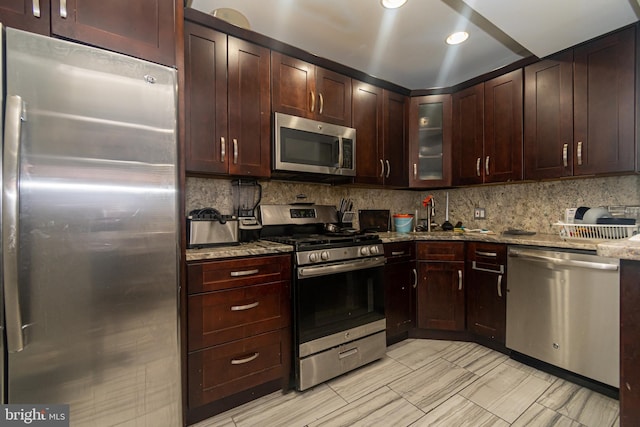 kitchen with light stone counters, a sink, backsplash, stainless steel appliances, and glass insert cabinets