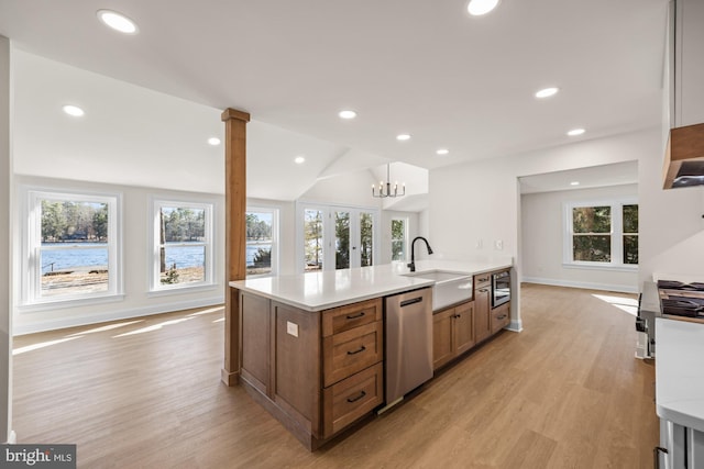 kitchen with brown cabinetry, an island with sink, a sink, stainless steel dishwasher, and light wood-type flooring