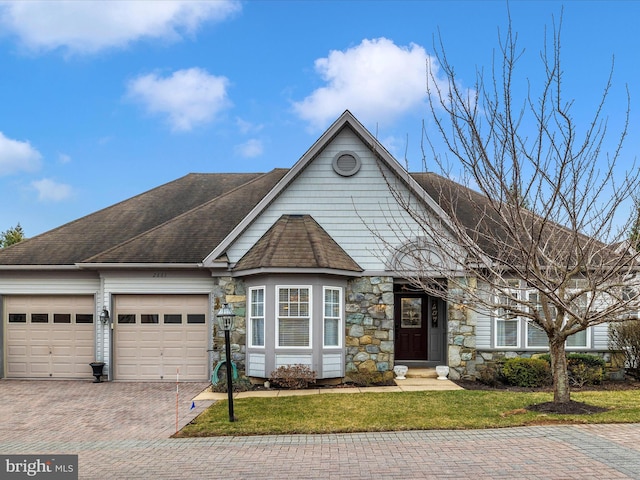 view of front facade with a shingled roof, a front lawn, a garage, stone siding, and decorative driveway