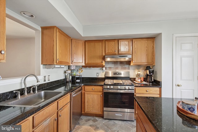 kitchen featuring under cabinet range hood, stone finish flooring, dark stone countertops, a sink, and appliances with stainless steel finishes