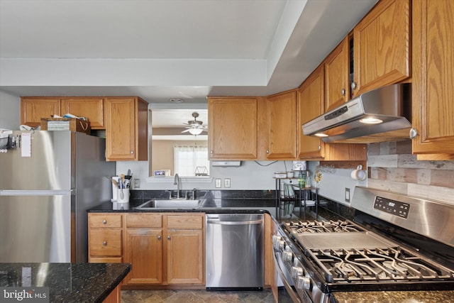 kitchen with dark stone countertops, brown cabinetry, a sink, under cabinet range hood, and appliances with stainless steel finishes