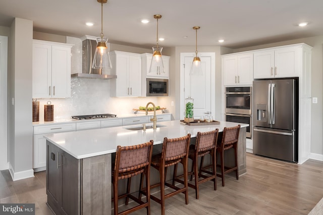 kitchen with a sink, white cabinets, stainless steel appliances, wall chimney exhaust hood, and a kitchen island with sink