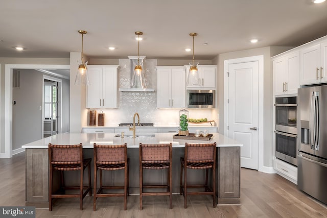 kitchen featuring a kitchen island with sink, stainless steel appliances, light countertops, white cabinets, and wall chimney exhaust hood