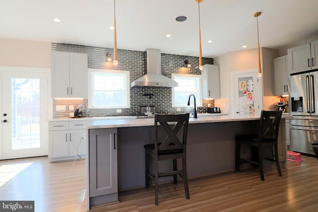 kitchen with wall chimney range hood, light countertops, light wood-type flooring, decorative backsplash, and stainless steel refrigerator with ice dispenser