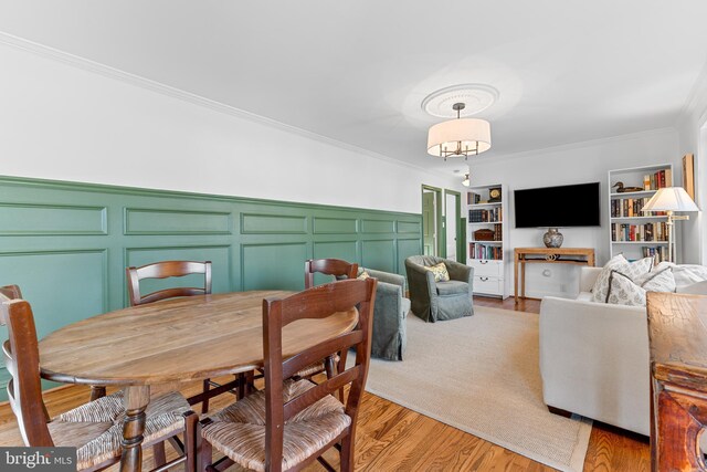 dining room with built in shelves, a wainscoted wall, crown molding, a decorative wall, and light wood-type flooring