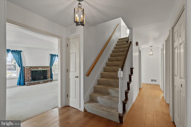 staircase featuring wood finished floors, visible vents, baseboards, a brick fireplace, and a notable chandelier