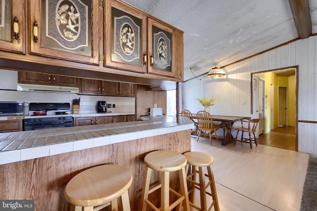 kitchen featuring stainless steel microwave, range with gas cooktop, exhaust hood, light wood finished floors, and tile counters