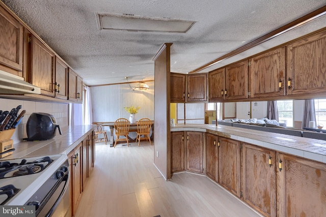 kitchen with light wood finished floors, gas range gas stove, tile counters, a textured ceiling, and brown cabinets