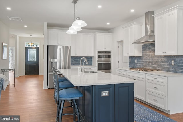 kitchen featuring a sink, wood finished floors, stainless steel appliances, white cabinets, and wall chimney range hood