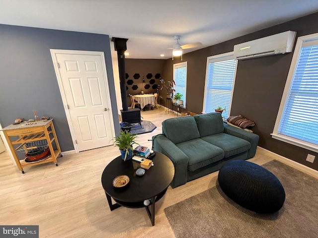 living room featuring baseboards, light wood-type flooring, a wood stove, a wall mounted AC, and a ceiling fan